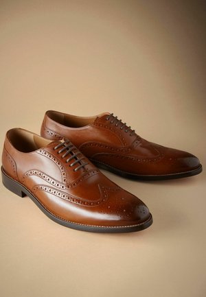 Chaussures à lacets en cuir marron avec un design à bout d'aile, des détails en brogue et des orteils arrondis. Elles disposent d'une semelle noire et de lacets fins.