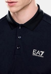 Camisa polo negra con cuello clásico, que presenta tres botones y un logo EA7 Emporio Armani bordado en blanco en el lado izquierdo del pecho.