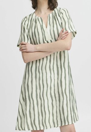 Kvinde med sammenlagte arme iført knælængde kjole med korte ærmer og lodrette, bølgede grønne striber på off-white baggrund.