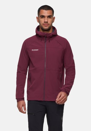 Homme portant une veste à capuche Mammut bordeaux fermée, se tenant devant un fond uni clair, avec un pantalon noir.