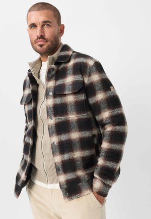 Mand iført sort, hvid og brun ternet knap-op jakke over beige lynlås sweater og cremefarvede kordfløjlsbukser mod hvid baggrund.