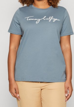 Person wearing a blue short-sleeve t-shirt with "Tommy Hilfiger" logo and beige pants, standing against a plain background.