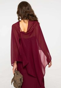 Shawl bordeaux trasparente con un design fluente. Presenta una schiena scoperta e una texture morbida. Una borsa con motivo è tenuta in mano.