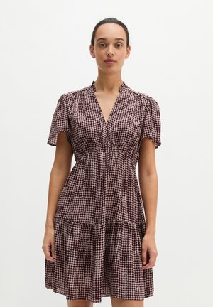 Donna in piedi che indossa un vestito a maniche corte, scollo a V e lunghezza al ginocchio con motivo a pois marrone e rosa chiaro su sfondo uniforme.