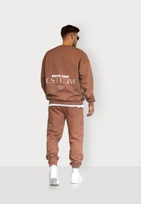 Brown sweatshirt and joggers set, featuring white printed text on the back, with ribbed cuffs and a relaxed fit, paired with white sneakers.