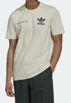 Uomo che indossa una t-shirt beige Adidas con la scritta "Mellow Ride Club" e pantaloncini neri, con la mano in tasca, su sfondo semplice.