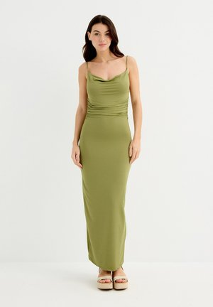 Vestido maxi verde oliva con cuello en cowl y lados fruncidos, confeccionado en un material elástico. Combinado con sandalias de cuña de plataforma.