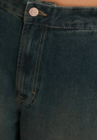 Tmavě modré džíny z denimu se měděným zapínáním, nabízející jemné švy a hladkou texturu. Detailní pohled na oblast pasu.