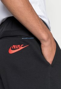 Svarta joggers i mjukt tyg med en broderad röd Nike-logga och en ljusblå detalj nära midjebandet.