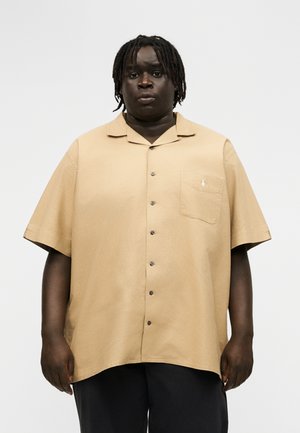 Μαύρο άτομο με κοντά dreadlocks που φοράει μπεζ πουκάμισο με κοντά μανίκια και τσέπη στο στήθος, μαζί με μαύρο παντελόνι, στέκεται μπροστά σε λευκό φόντο.