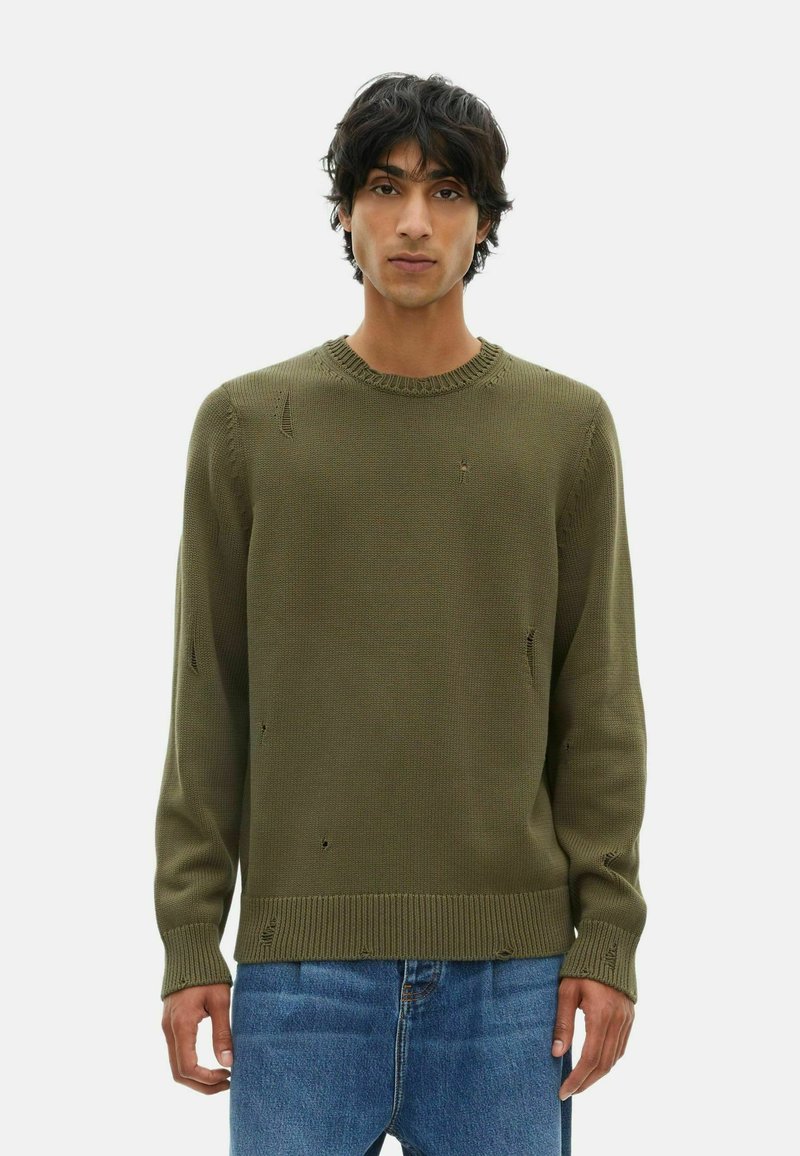 Joven vestido con un suéter de punto color verde olivo con agujeros intencionales y jeans de mezclilla azules, de pie contra un fondo blanco.