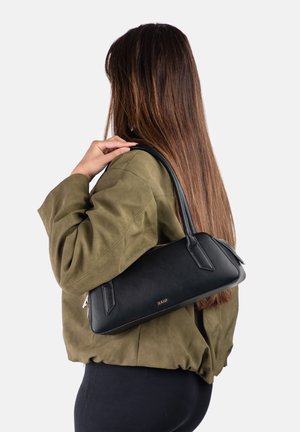 Femme aux longs cheveux bruns portant une veste vert olive, portant un sac à bandoulière noir avec des détails de fermeture éclair dorée sur son épaule droite.