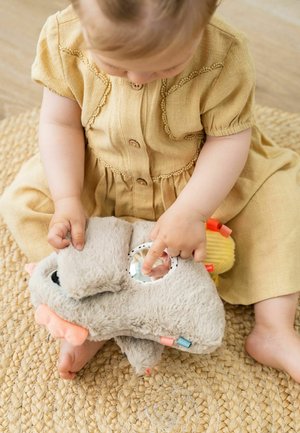 Grau verschmustes Spielzeug, das ein Tier ähnelt, mit einem glänzenden runden Spiegel, weichem Material und bunten Stoffakzenten. Ein Kind interagiert damit auf einem gewebten Teppich.