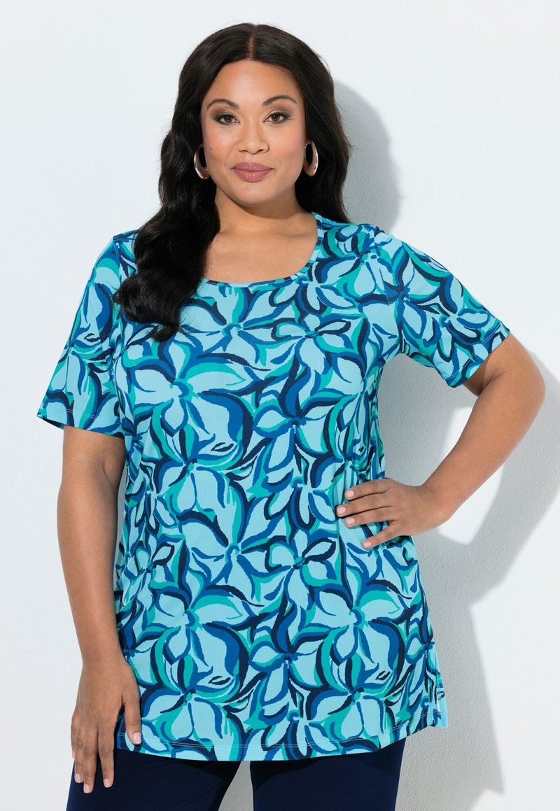 Mujer con cabello largo y oscuro, llevando pendientes de aro y una blusa de manga corta con estampado floral azul, de pie con una mano en la cadera.