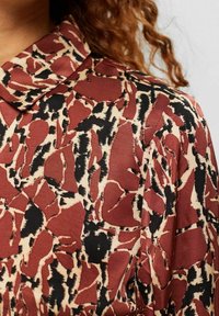 Blusa astratta rossa e nera con colletto, caratterizzata da una texture liscia e un design fluido. Il tessuto sembra leggero.