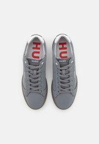 Grå lær sneakers med rund tå, flade snørebånd og polstret krave. Har rød "HUGO" logo på tungen og siden. Gummisål.