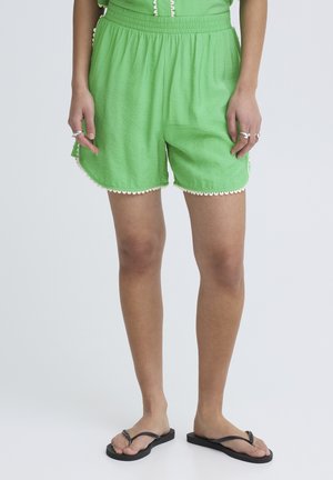 Persona con pantalones cortos verdes brillantes con cintura elástica y bordes ondulados, chanclas negras y anillos plateados en los dedos.