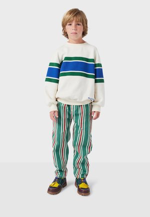 Sweatshirt blanc avec des rayures horizontales vertes et bleues, associé à un pantalon rayé vert avec des lignes rouges et blanches. Chaussures marron avec des accents jaunes.
