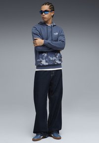 Blå hoodie med camouflage mønster i bunden, parret med løse mørke denim bukser og blå sneakers. Accessories med blå solbriller.
