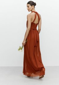Maxime dress in ruskea, valmistettu kevyistä kankaista. Ominaisuuksia ovat halterneck-malli, solmittava vyötärö ja läpikuultava päällyste.