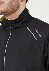 Veste noire zippée avec col haut, présentant des accents gris réfléchissants et une doublure intérieure jaune. Conçue en matériau lisse et léger.