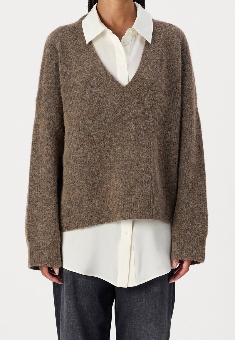 En person som bär en brun V-ringad ulltröja över en vit skjorta i knappar, ihop med mörkgrå byxor mot en neutral bakgrund.