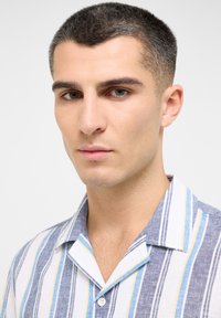 Camisa a rayas con líneas azules, blancas y amarillas, fabricada en un tejido ligero, con cuello con muesca y cierre de botones. Vista de cerca.