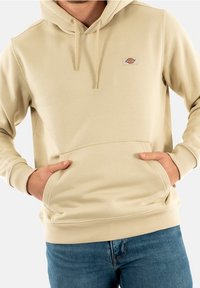 Felpa con cappuccio beige realizzata in un tessuto morbido, dotata di tasca frontale a marsupio e polsini a costine. Piccola toppa con logo sul petto.