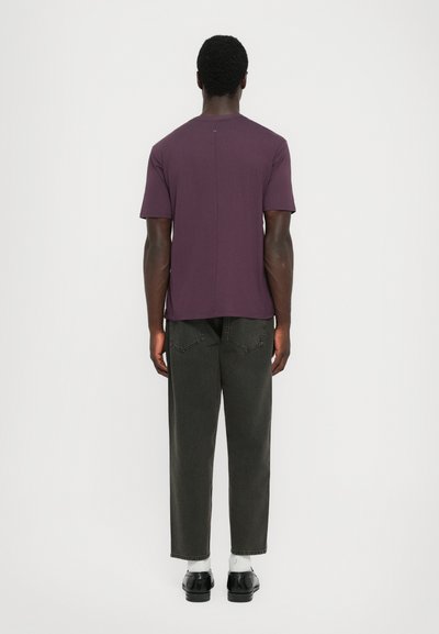 Man standing facing away wearing a short-sleeve purple shirt, black jeans, white socks, and black dress shoes on a plain white background.