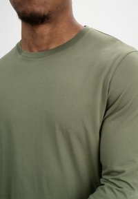 Olivengrünes langärmliges T-Shirt aus weichem Baumwollstoff. Es hat einen Rundhalsausschnitt und eine glatte Textur mit einer schlanken Passform.