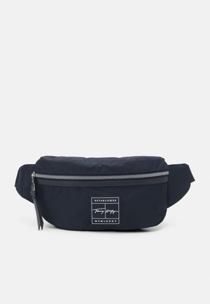 Sac banane rectangulaire noir avec fermeture éclair argentée et patch logo blanc indiquant "Established Tommy Hilfiger MCMLXXXV."
