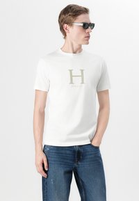 Hvitt bomull T-skjorte med et stort beige "H"-logo og "HACKETT"-tekst under. Kombinert med blå jeans og mørke solbriller.