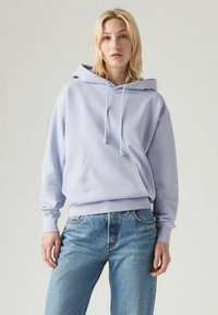 Felpa viola chiaro con cappuccio, stringhe, tasca a marsupio, polsini e orlo a costine. Abbinata a jeans di denim blu chiaro.