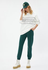 Biała bluza z napisem "MONTECARLO ATHLETIC VIBES", zestawiona z zielonymi spodniami dresowymi i białymi sznurowanymi butami. Ciemnozielona czapka z logo "Y".
