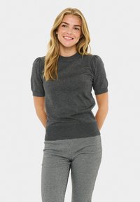 Femeie zâmbitoare cu păr lung blond, purtând un top tricotat cu mâneci scurte de culoare gri închis și pantaloni în carouri alb-negru.