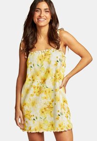 Florale zomerjurk met gele bloemen op een witte achtergrond, spaghettibandjes en decoratieve kantafwerking aan de zoom. Lichtgewicht stof.