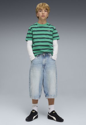 Camiseta de rayas verdes, con mangas largas blancas debajo. Pantalones cortos sueltos de mezclilla azul, zapatillas negras con detalles dorados, calcetines blancos. Fondo neutro.