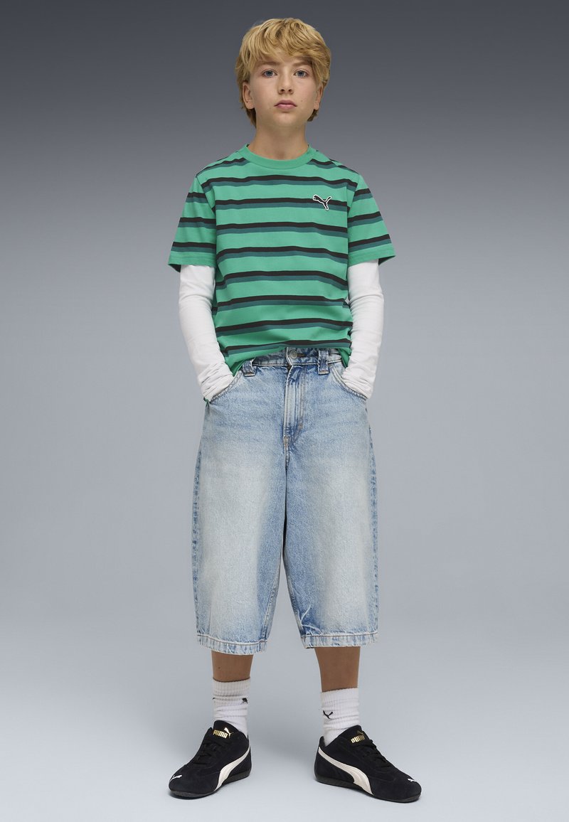 Zielona koszulka w paski, pod spodem białe długie rękawy. Luźne niebieskie szorty z denimu, czarne tenisówki ze złotymi akcentami, białe skarpetki. Neutralne tło.