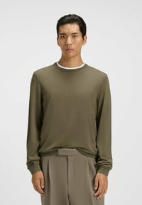 Olivegrünes Langarmshirt mit Rundhalsausschnitt, leichter Textur und kontrastierendem weißen Kragen. Kombiniert mit beigen Hose.