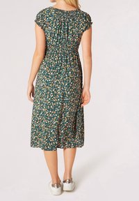 Tummaanvihreä kukkamekko, jossa on lyhyet röyhelöhihat, joustava vyötärö ja keskipitkä helma. Värikkäitä kukkakuvioita kauttaaltaan. Päälle valkoiset tennarit.