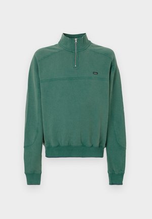 Grønn hettegenser med høy krage og halv glidelås, med en avslappet passform, ribbestrikkede mansjetter og en liten logo på brystet.