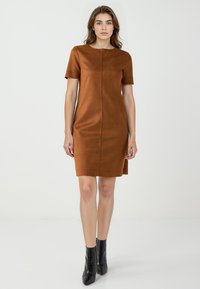 Braunes Kurzarmkleid aus weichem, strukturiertem Stoff. Hat einen geraden Schnitt und eine Naht vorne. Getragen mit schwarzen Stiefeletten.