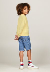 Felpa gialla, pantaloni corti in denim blu, sneakers bianche con dettagli rossi e blu navy, e calze a righe. Capelli ricci visibili. Vista dorsale di un bambino.