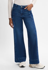 Weite, dunkelblaue Denim-Jeans mit hoher Taille, sichtbaren Nähten und Vorderaschen. Kombiniert mit hellen Sportschuhen.