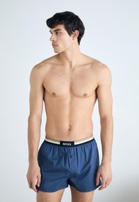 Boxers pour homme en bleu, fabriqués en tissu léger, avec une ceinture élastique noire et dorée portant le logo "BOSS" et une fermeture à un bouton.