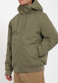 Olijfgroene hoody met rits, ribgebreide manchetten en zoom, zijzakken en een klein logo op de borst. Glad textuur van de stof.
