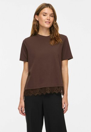 Femme aux cheveux châtain clair portant un haut marron à manches courtes avec bordure en dentelle et un pantalon noir, debout devant un fond uni.
