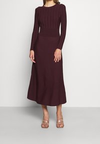 Robe en maille bordeaux à manches longues, texture côtelée sur le corsage, taille cintrée et ourlet évasé. Portée avec des talons transparents.