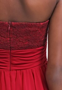 Robe rouge avec un corsage en dentelle texturée et une jupe en tissu froncé. Le dos présente une fermeture éclair invisible et des détails plissés.