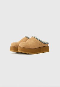 Brune suede klomper med en teksturert, fargerik vevd kant og en lys brun plattformsåle. Har rund tå og et slip-on design.